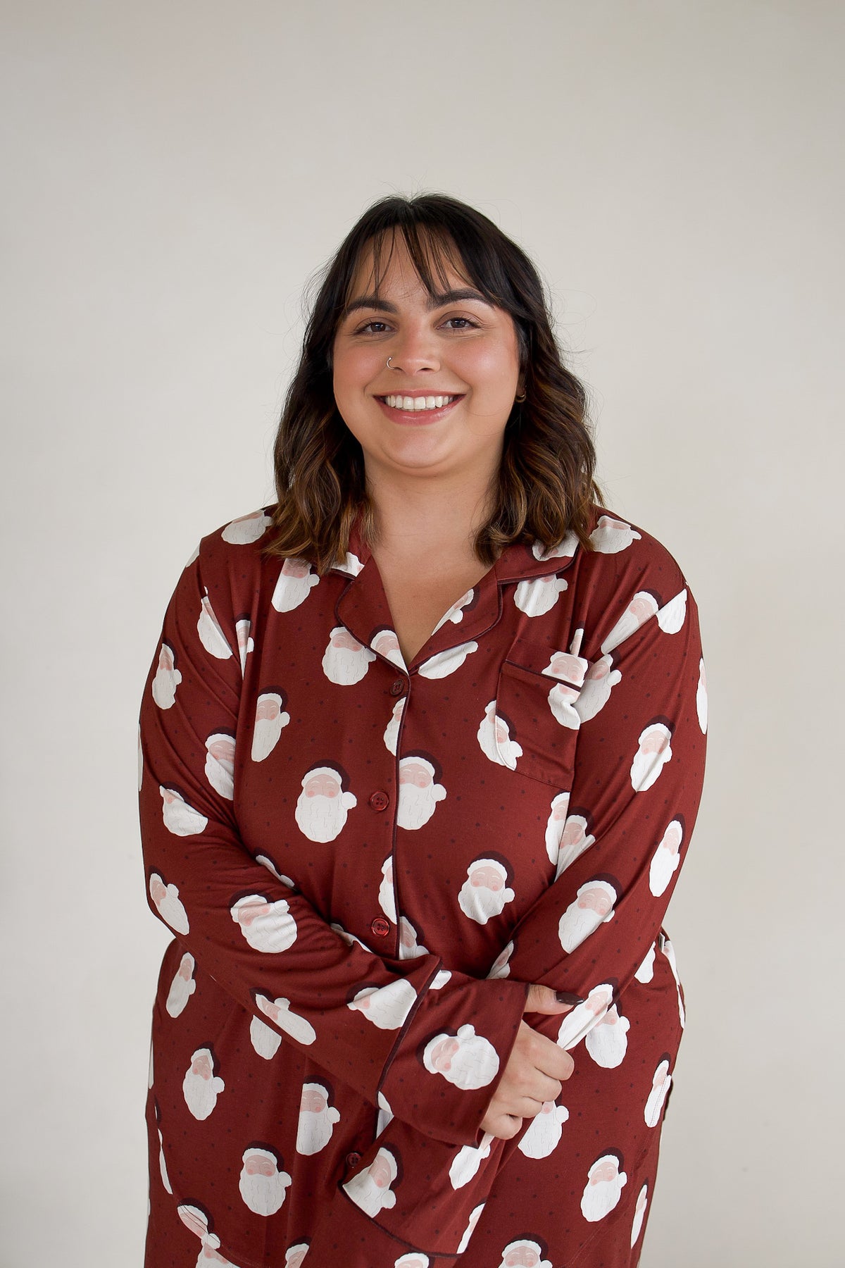 A smiling woman wears a maroon long-sleeved Women’s Bamboo Button Up Lounge Set | Vintage Santa featuring a repeating pattern of Santa Claus faces.