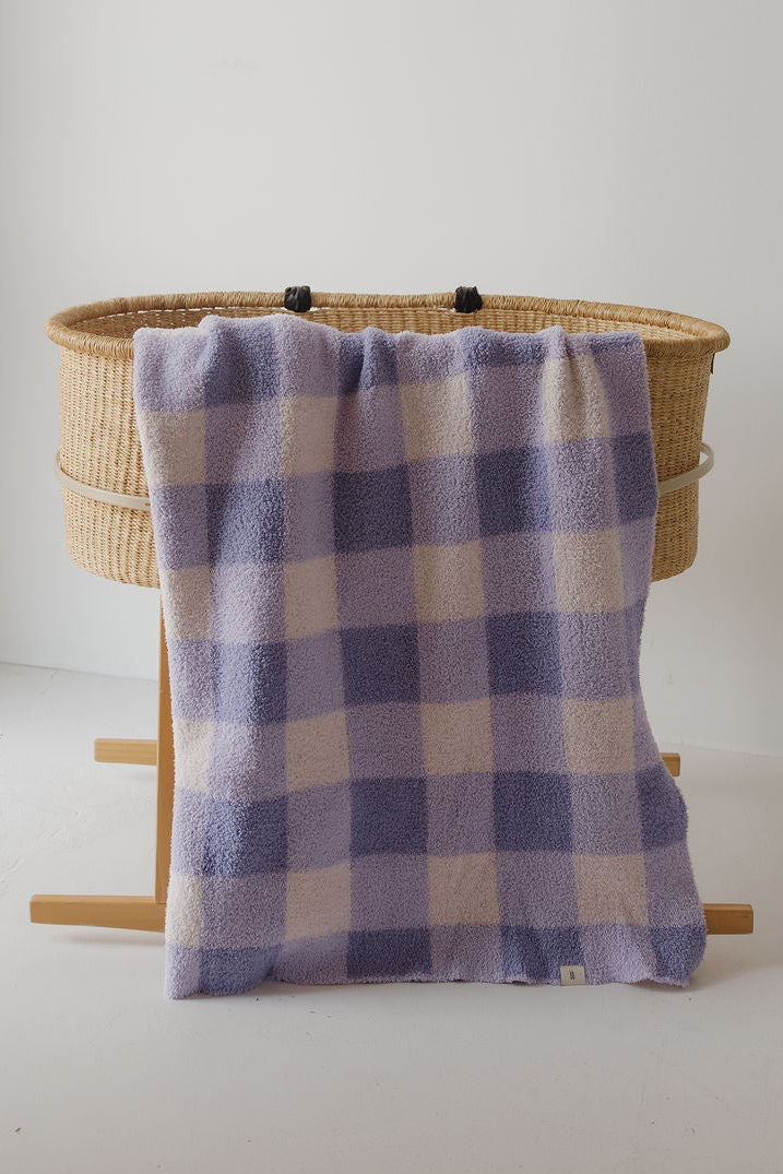 A woven bassinet with wooden legs stands against a white wall, draped with the forever french baby Plush Blanket | Marilyn—a soft lavender and cream checkered, OEKO-TEX certified baby blanket.
