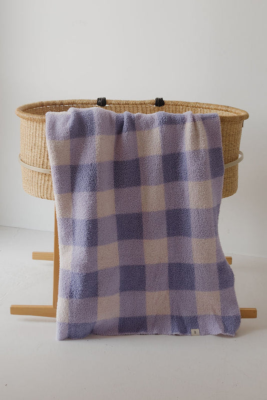 A woven bassinet with wooden legs stands against a white wall, draped with the forever french baby Plush Blanket | Marilyn—a soft lavender and cream checkered, OEKO-TEX certified baby blanket.