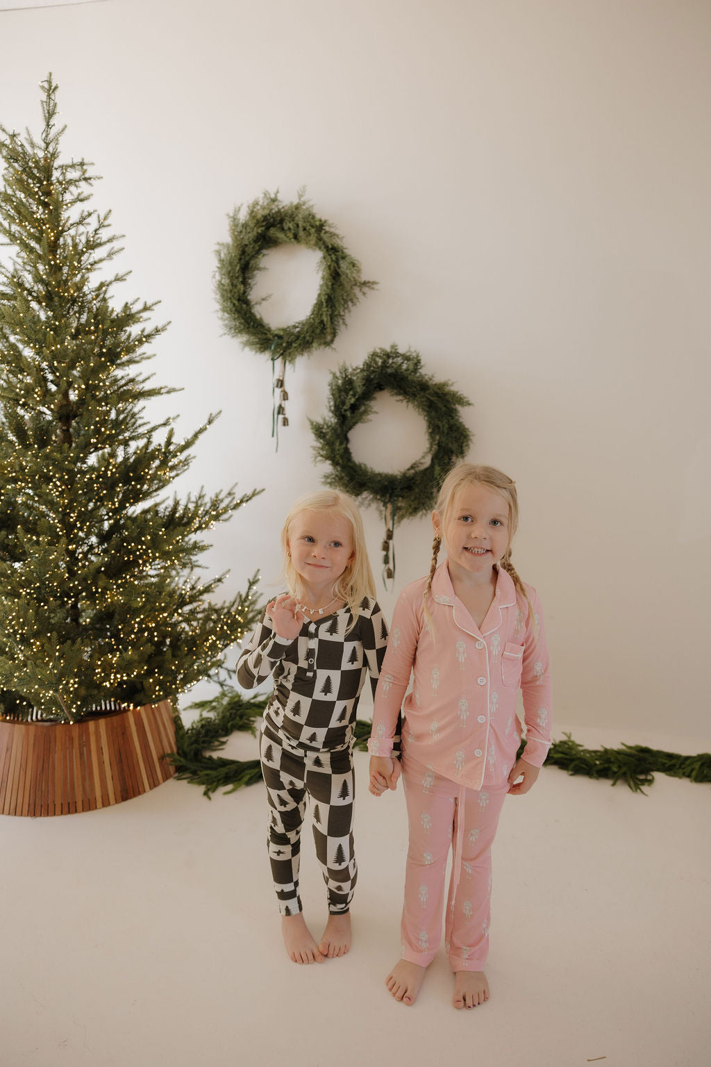 Two young girls in forever french baby Bamboo Two Piece Pajamas—one wearing the Christmas Tree Checkerboard pattern, the other in pink—stand barefoot and hand-in-hand before a decorated Christmas tree and three wreaths on a white wall.