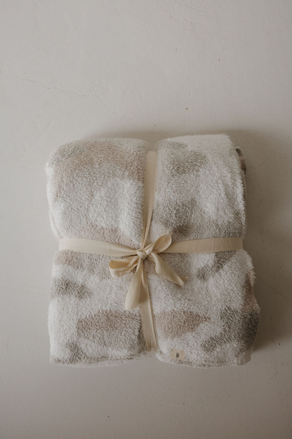 The forever french baby Plush Blanket | Camo 2.0 features a soft, folded microfiber design in beige and white, tied with a cream ribbon and displayed on a light surface.