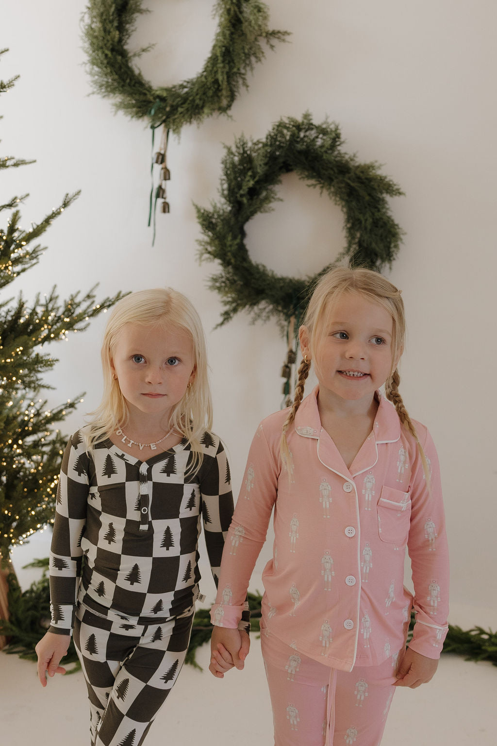 Two young girls hold hands by a decorated Christmas tree and two wreaths. One wears forever french baby Bamboo Two Piece Pajamas in Christmas Tree Checkerboard; the other wears pink kids’ pajamas with a festive holiday print.