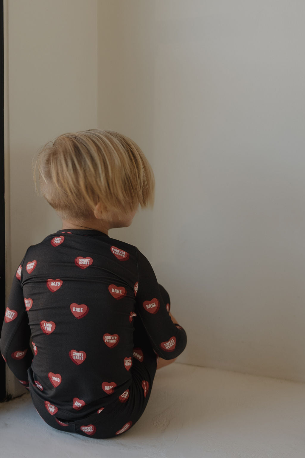A young child with blonde hair sits on the floor facing a light wall, wearing forever french baby's Bamboo Two Piece Pajamas | Love Notes – Charcoal—black hypoallergenic sleepwear adorned with red hearts and playful messages.