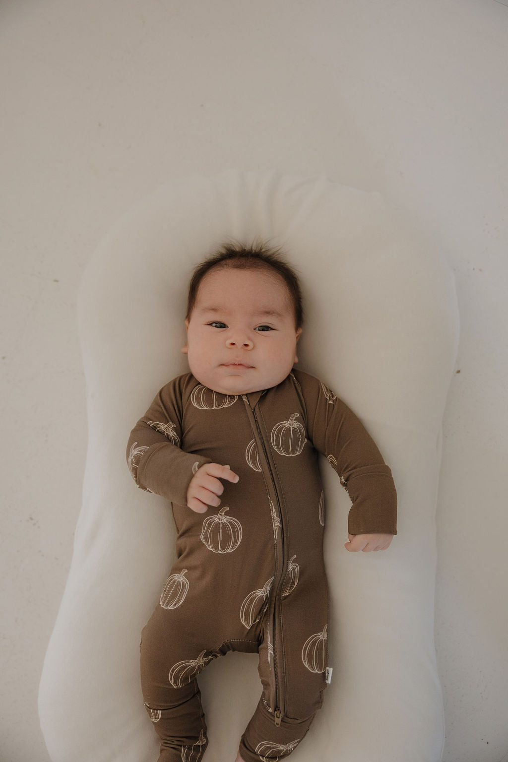 A baby in forever french baby's Bamboo Zip Pajamas | Brown Pumpkin lies calmly on a white, cushioned surface, gazing upward.