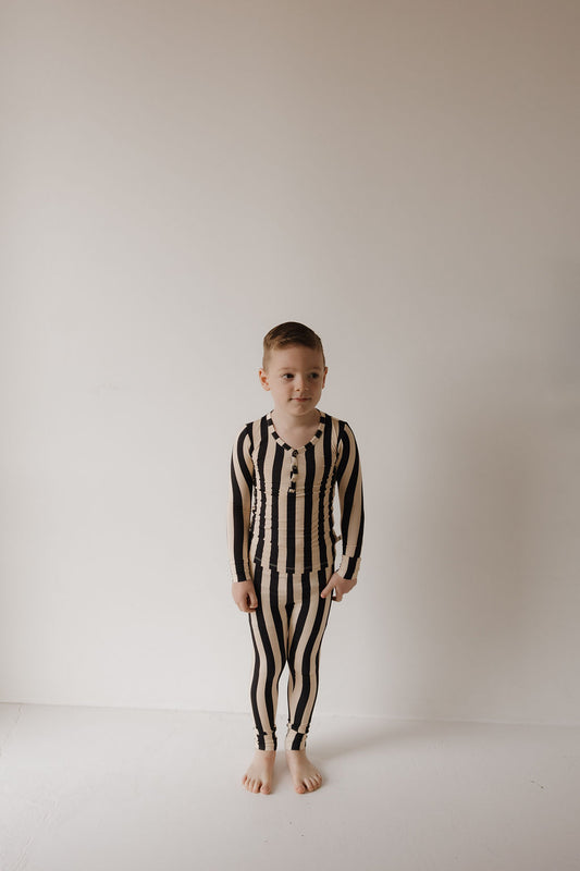 A young boy stands barefoot against a white wall, wearing Minty Cherries’ Bamboo Two Piece Pajamas in the Midnight Stripe pattern. He looks off to the side with a neutral expression.