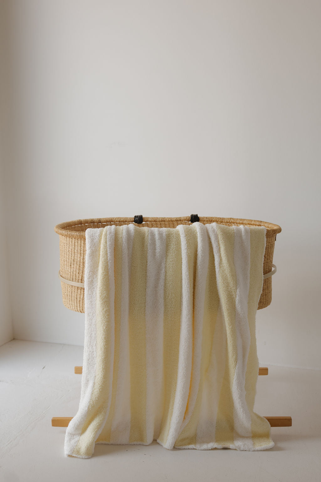 A wicker bassinet on wooden legs with the Plush Blanket | Sunny Stripe by forever french baby—an Oeko-Tex certified, soft baby blanket in striped yellow and white—draped over the side, set against a plain white wall and floor.