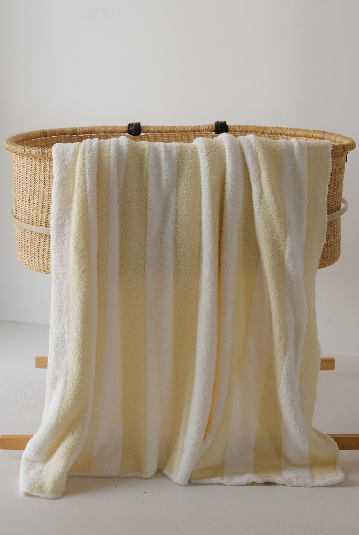 A Plush Blanket | Sunny Stripe by forever french baby, featuring soft white and yellow stripes, is draped over the side of a woven bassinet against a plain white background.