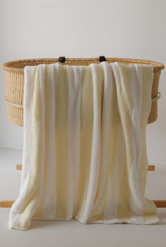 A Plush Blanket | Sunny Stripe by forever french baby, featuring soft white and yellow stripes, is draped over the side of a woven bassinet against a plain white background.