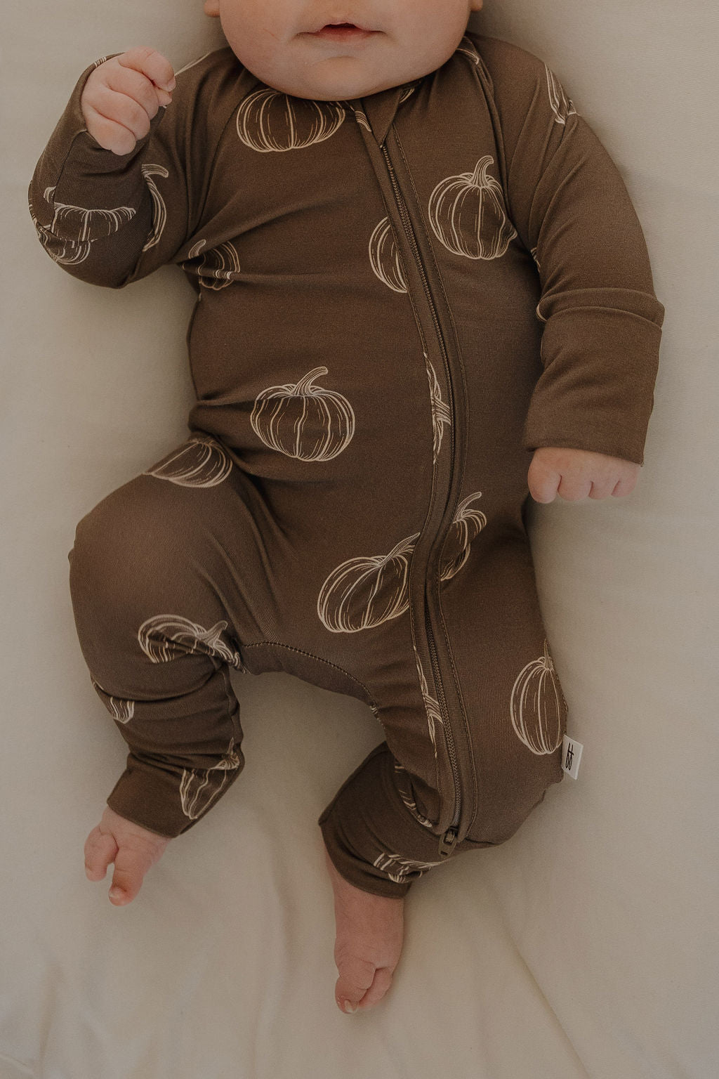 A baby lies on a bed wearing forever french baby's Bamboo Zip Pajamas in Brown Pumpkin—cozy and breathable sleepwear.