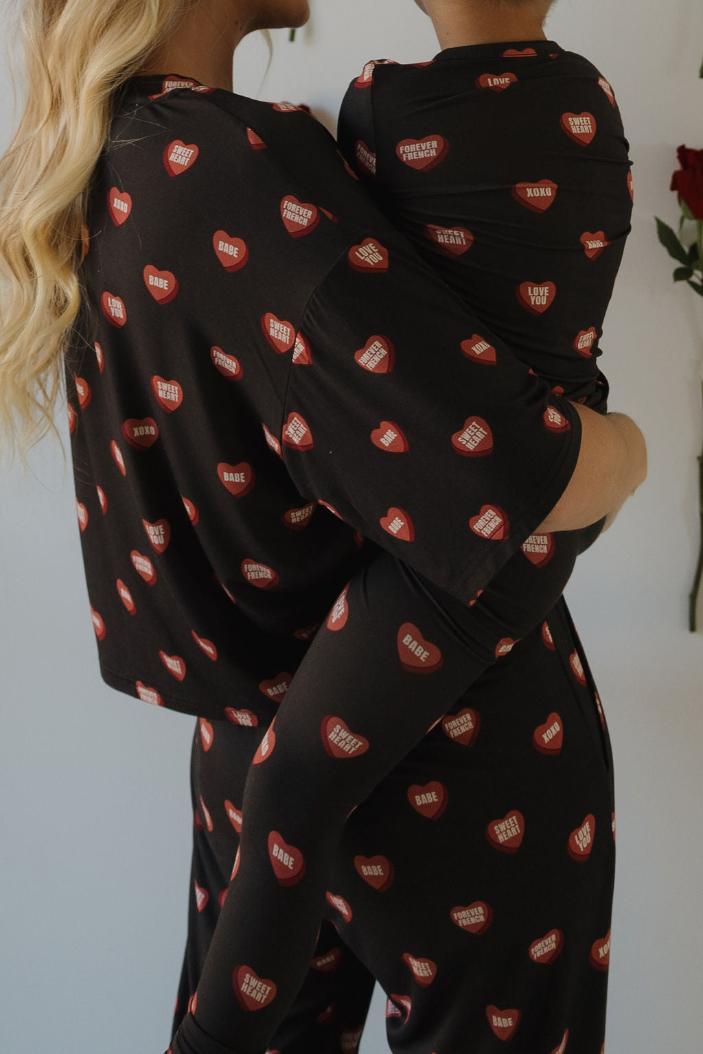 A woman holds a child, both wearing the forever french baby Women's Boxy Tee Lounge Set | Love Notes - Charcoal, featuring red hearts and sweet phrases. They face each other against a softly lit background with roses visible.