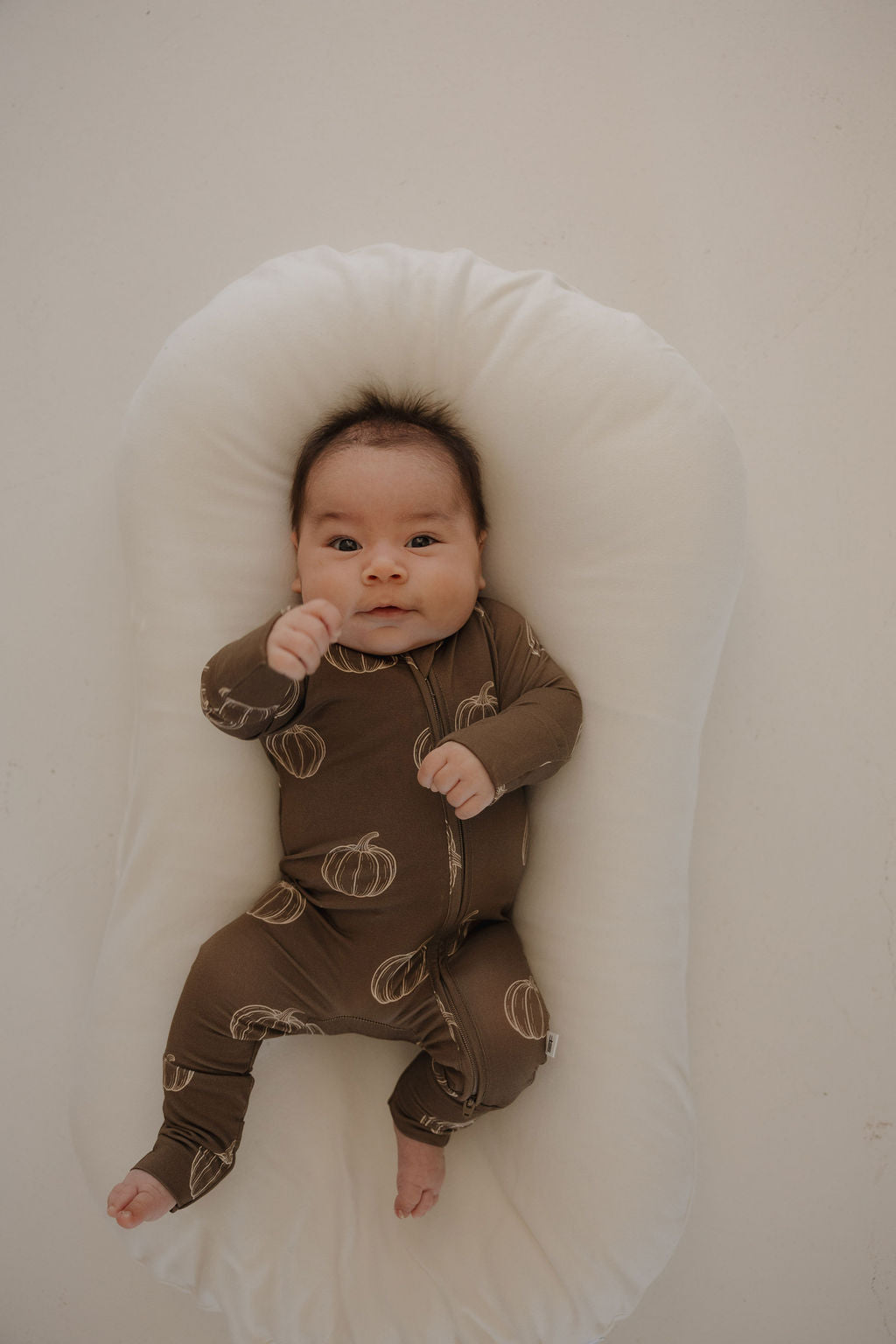 A baby in forever french baby Bamboo Zip Pajamas | Brown Pumpkin lies on a white, cushioned baby lounger, looking up with one hand slightly raised.