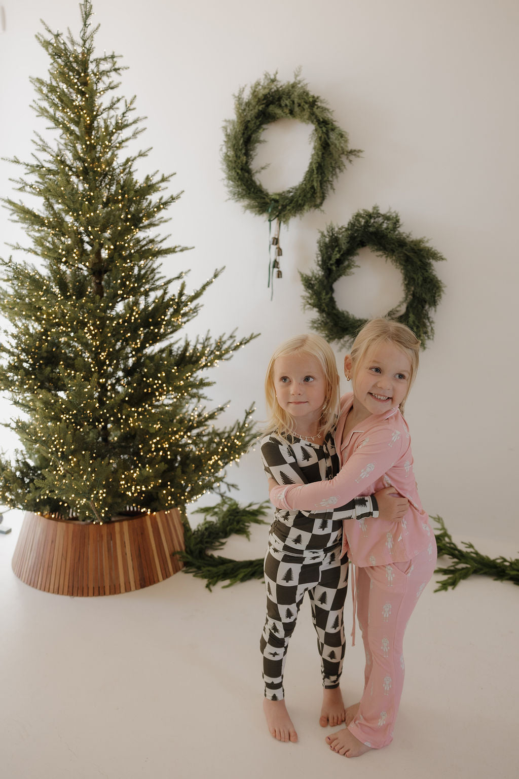 Two young children in forever french baby Bamboo Two Piece Pajamas | Christmas Tree Checkerboard hug and smile by a lit Christmas tree. Three green wreaths hang on the white wall, with greenery scattered on the floor.
