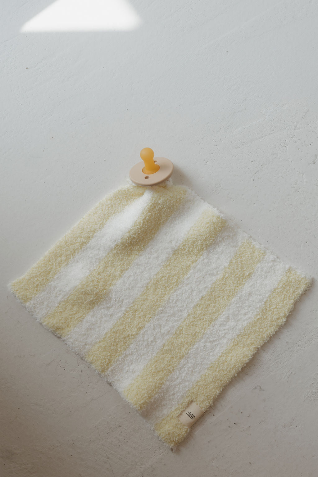A yellow and white striped soft cloth, the Lovey | Sunny Stripe by forever french baby, lies flat on a light surface with a tan and yellow pacifier resting on top near one corner.