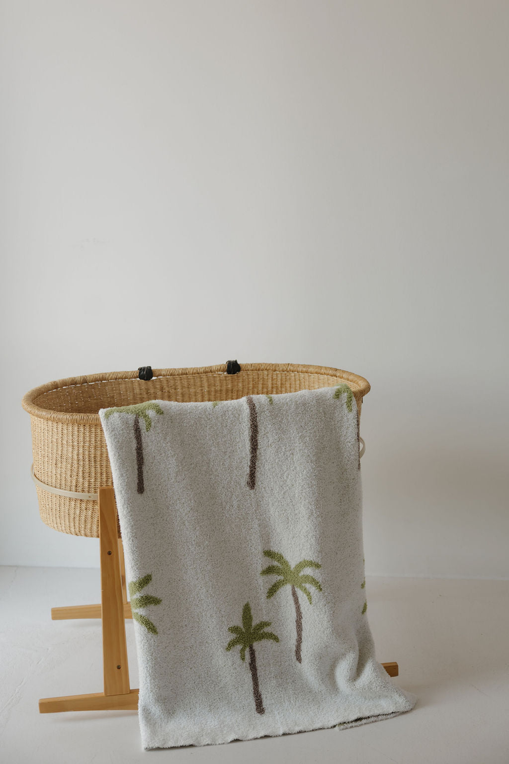 A woven bassinet with a wooden stand is draped with the forever french baby Plush Blanket | Palm—an Oeko-Tex certified, soft white blanket adorned with green palm tree designs—set against a plain white wall and floor.