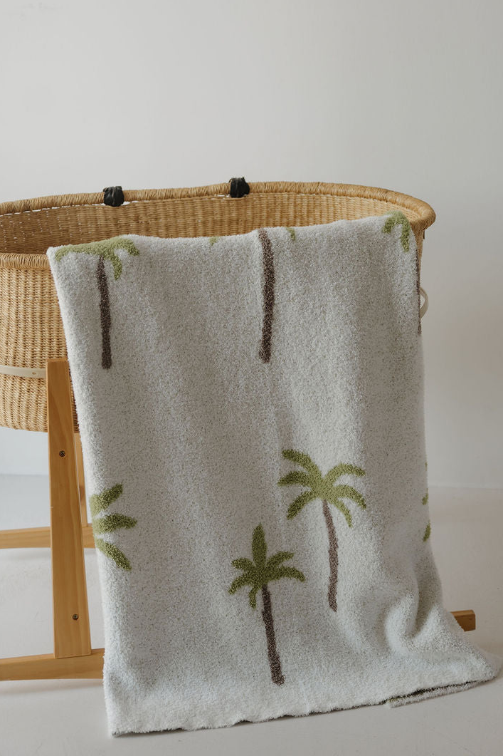 The forever french baby Plush Blanket | Palm, featuring a white textured design with green and brown palm tree patterns and Oeko-Tex certification, is draped over the side of a woven bassinet on a wooden stand, set against a plain white wall.