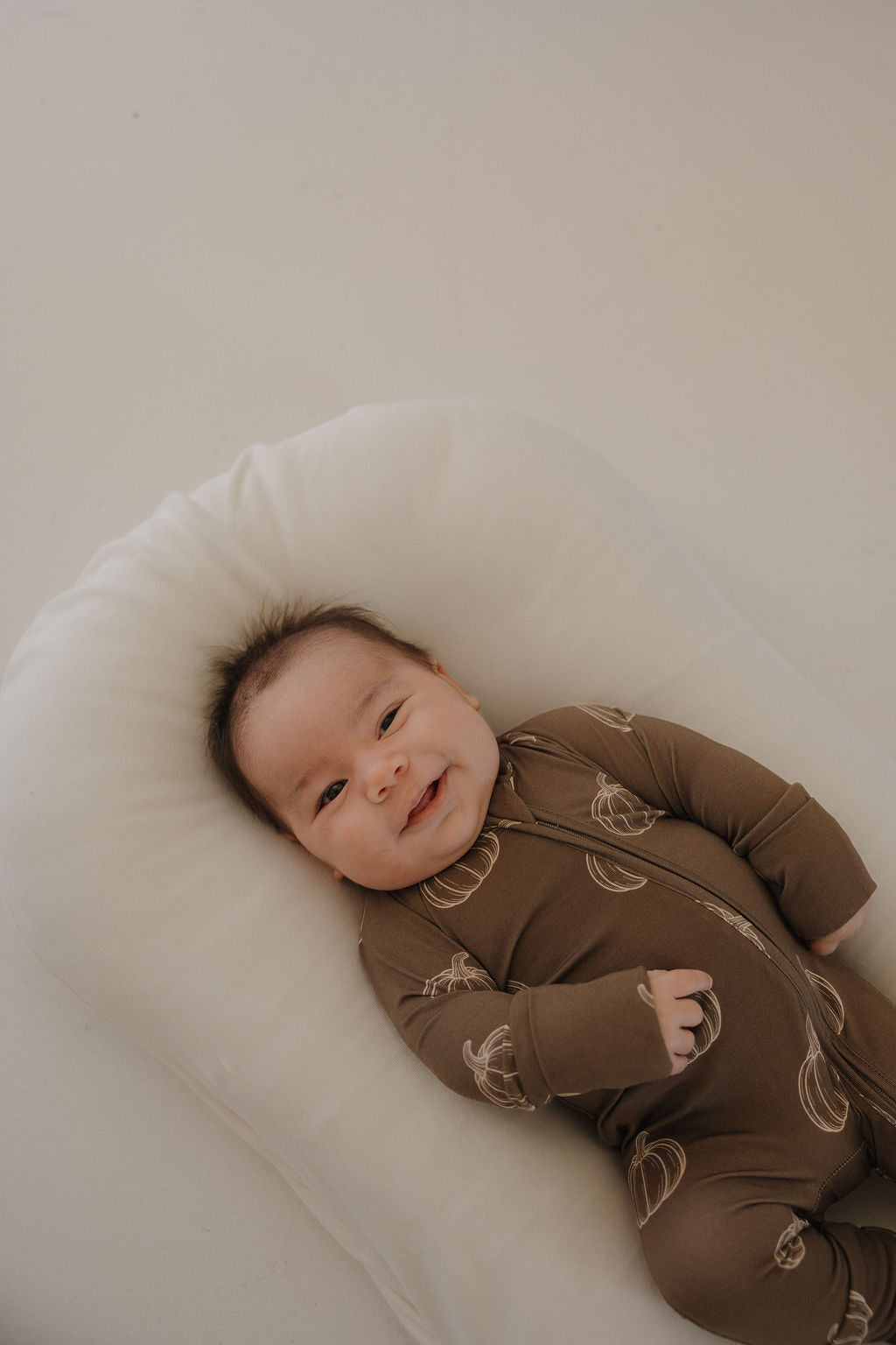 A smiling baby lies on a white cushion, relaxed and content, wearing forever french baby's Bamboo Zip Pajamas in Brown Pumpkin with a light pattern, against a neutral background.