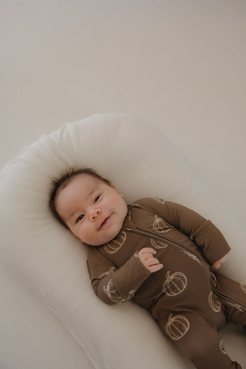 A baby in forever french baby's Bamboo Zip Pajamas | Brown Pumpkin, a breathable brown onesie with pumpkin designs, lies on a white cushioned pillow, looking up and smiling softly.