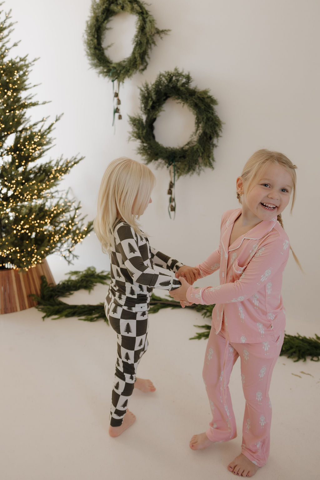Two young children in forever french baby's Bamboo Two Piece Pajamas | Christmas Tree Checkerboard hold hands and smile, standing in a festively decorated room with a lit Christmas tree and green wreaths on a white wall.
