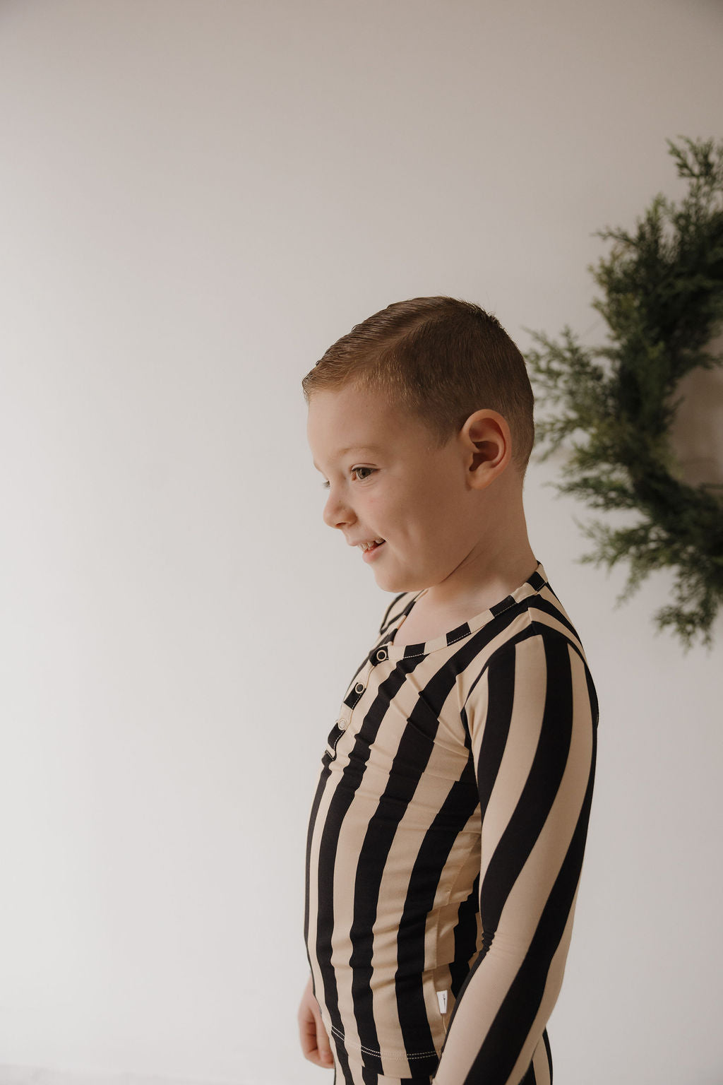 A young boy with short hair stands sideways, smiling in Minty Cherries' Bamboo Two Piece Pajamas | Midnight Stripe. A green wreath decorates the white wall behind him.