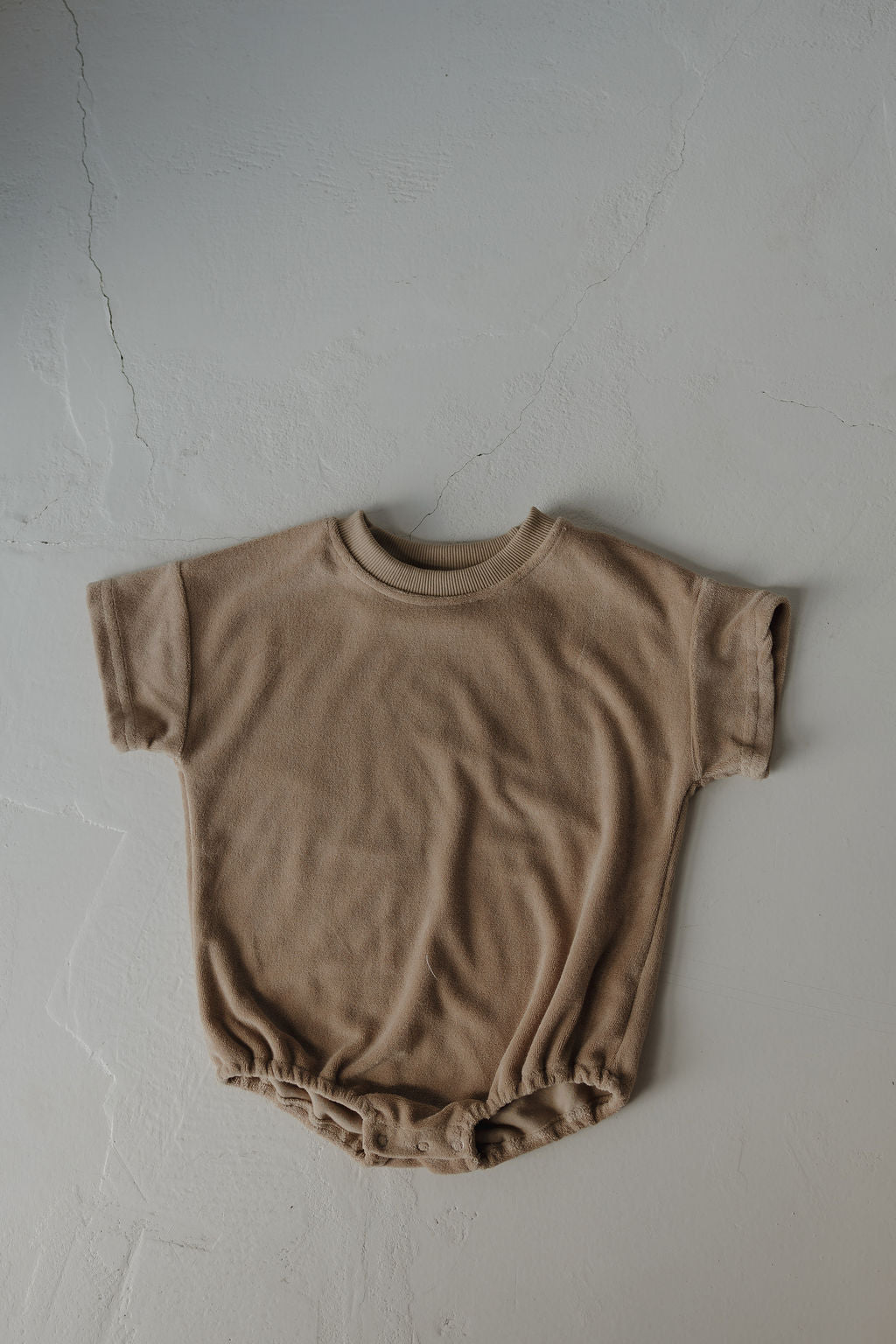 The forever french baby Terry T-Shirt Romper | Tan is a beige, short-sleeve terry onesie with a snap closure, shown laid flat on a textured light gray surface. Suitable for newborns to 24 months.