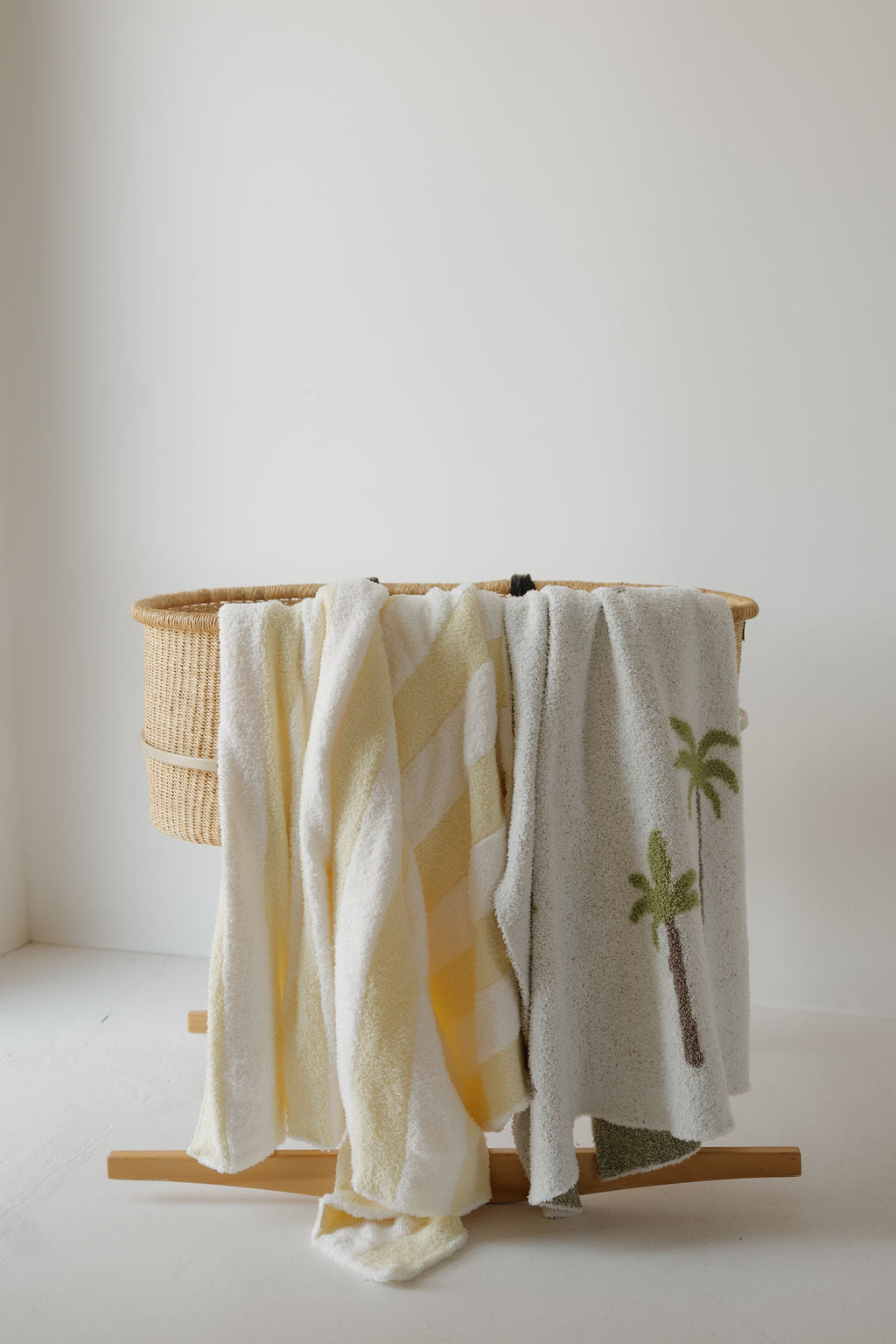 A woven bassinet on a wooden stand has three towels draped over it: a white towel, a yellow-and-white striped towel, and a light gray towel with palm tree designs. Beneath the towels rests the forever french baby Plush Blanket | Palm, which is Oeko-Tex certified for comfort and safety. The background is minimal and white.