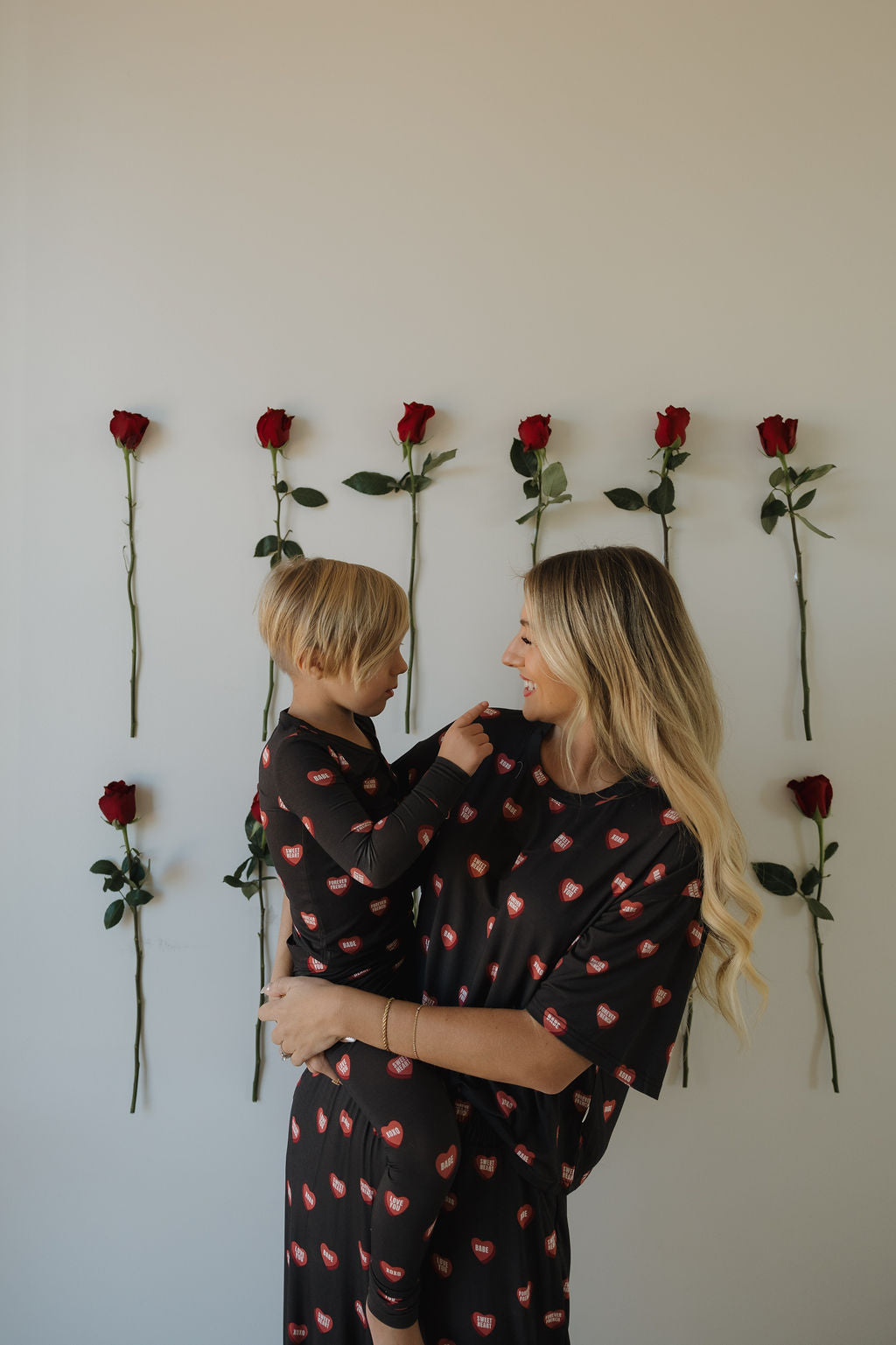 A woman and her child, both smiling in matching forever french baby Women's Boxy Tee Lounge Set | Love Notes - Charcoal outfits with red heart prints, stand before a plain wall decorated with vertical rows of red roses.