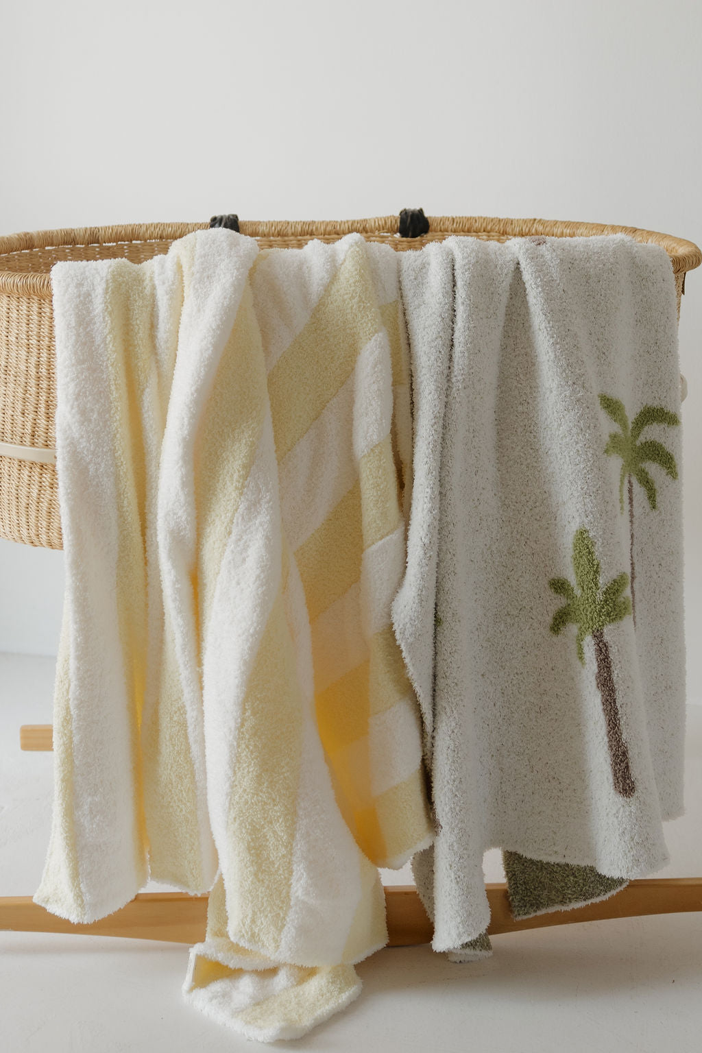 Three soft towels hang over the edge of a woven basket—one is white, one is yellow and white striped, and one is gray with embroidered green palm trees. Nestled beside them is the forever french baby Plush Blanket | Sunny Stripe, Oeko-Tex certified for added comfort and peace of mind.