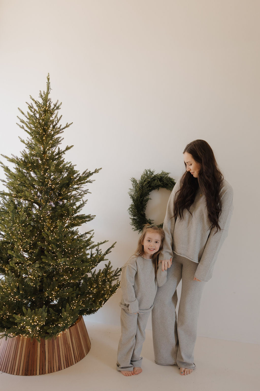 A woman and a young girl wearing forever french baby’s Knit Pant Set in Grey stand barefoot by a decorated Christmas tree and wall wreath, sharing warm smiles.