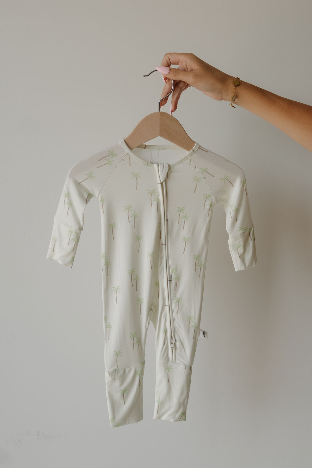 A hand holds a wooden hanger with the forever french baby Bamboo Zip Pajamas | Palm—a white baby onesie adorned with a light green palm tree pattern—displayed against a plain light wall; perfect for parents seeking hypoallergenic baby clothing that’s as stylish as it is gentle on delicate skin.