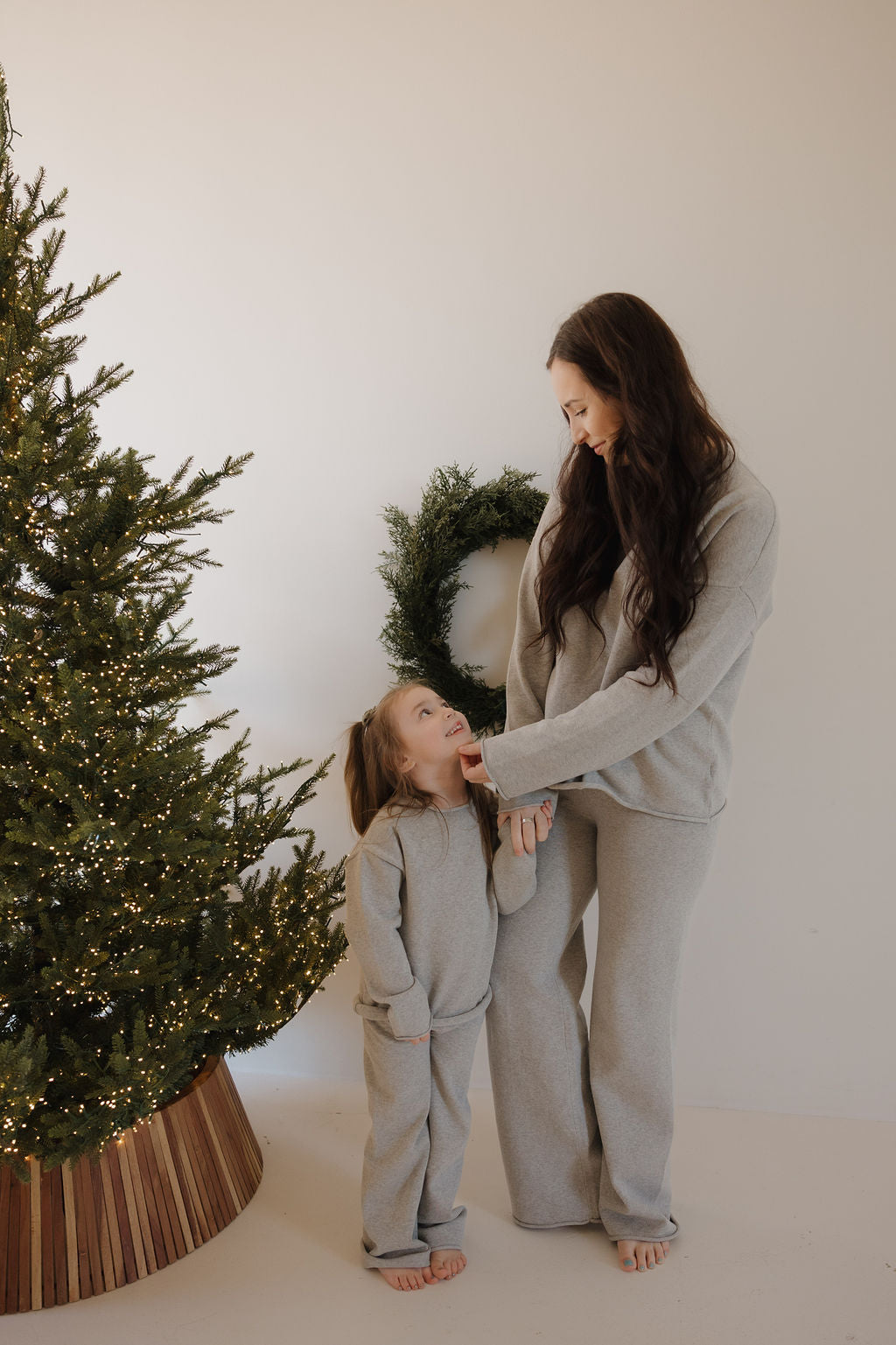 A woman and a young girl smile at each other, standing barefoot by a decorated Christmas tree. They wear matching forever french baby Knit Pant Set | Grey loungewear, made from 100% cotton. A green wreath hangs on the white wall behind them.