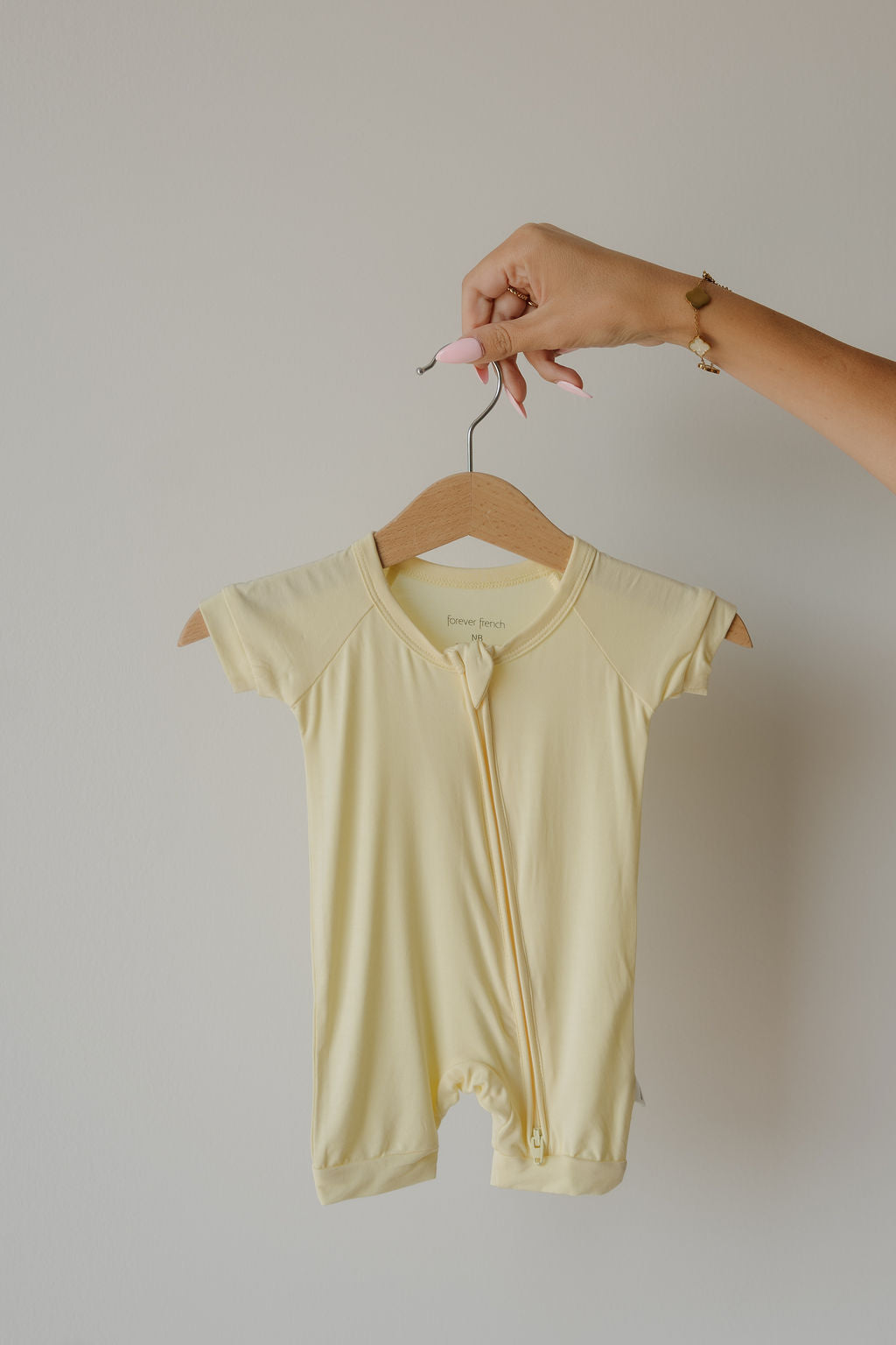 A hand holds a hanger with the forever french baby Shortie Bamboo Zip | Sunny, a short-sleeve, pale yellow romper, against a plain light-colored wall—showcasing the comfort and hypoallergenic softness of premium bamboo baby clothing.
