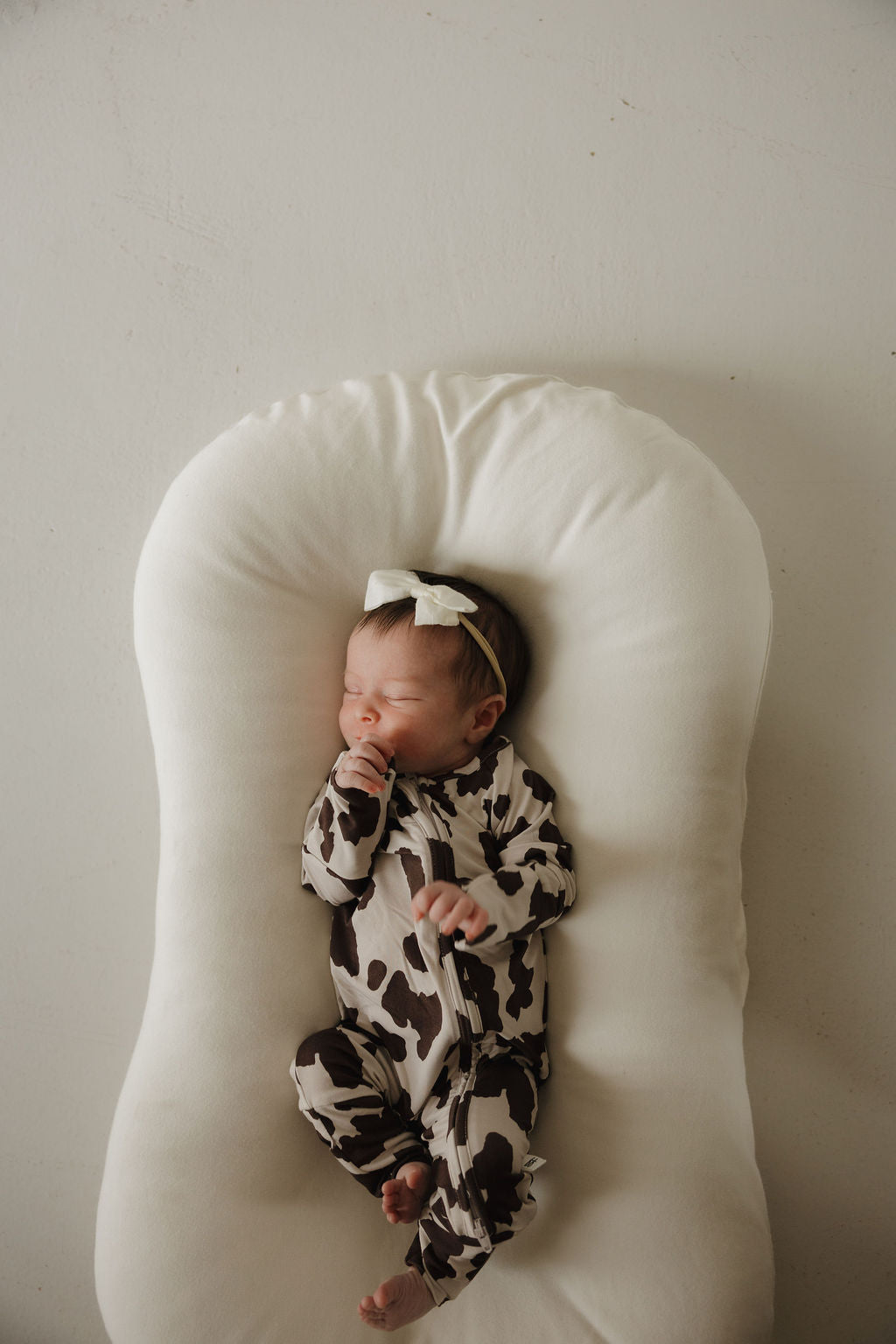 A newborn in forever french baby’s Bamboo Zip Pajamas | Moo! sleeps peacefully on a soft, white lounger, sporting a cow-print onesie and white bow headband against a light background.