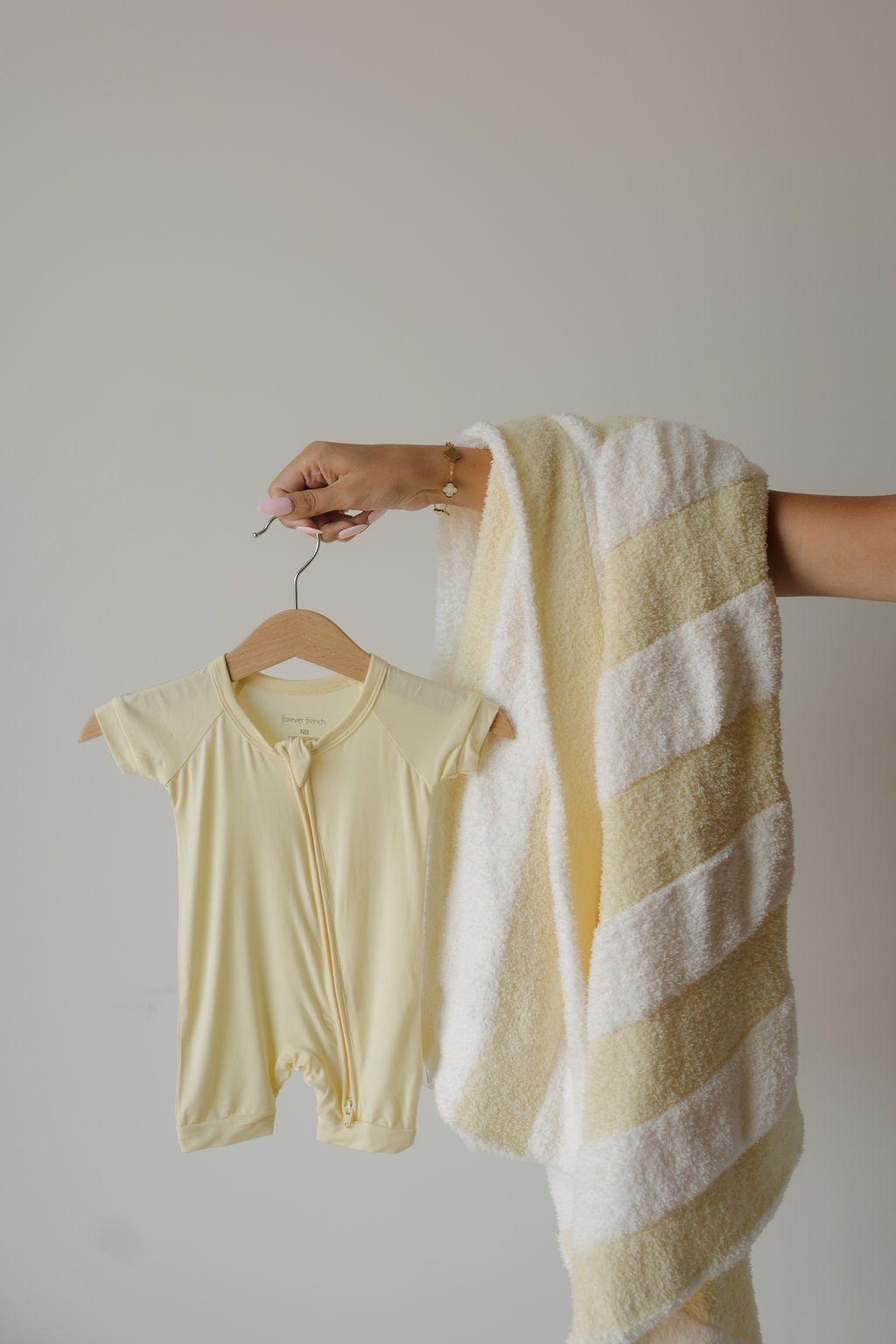 A hand holds the forever french baby Shortie Bamboo Zip | Sunny—a light yellow bamboo romper—on a hanger, alongside a yellow-and-white striped towel, against a plain, light background, highlighting the appeal of soft, hypoallergenic baby clothing.