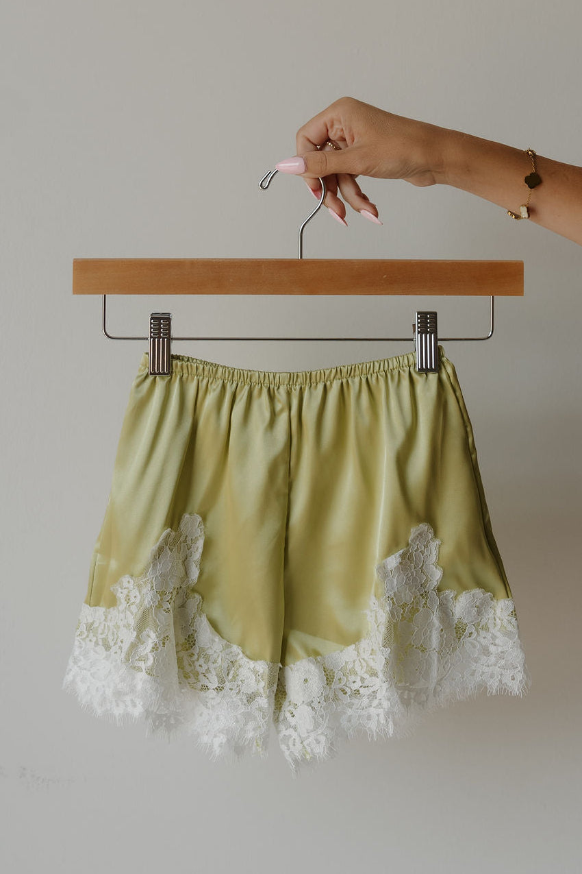 A hand holds a hanger with forever french baby’s Girl’s Satin Shorts | Prickly Pear—light green, lightweight shorts featuring delicate white lace trim at the hem—against a plain light background.