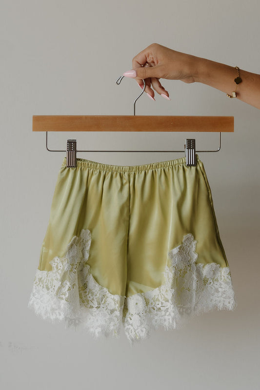 A hand holds a hanger with forever french baby’s Girl’s Satin Shorts | Prickly Pear—light green, lightweight shorts featuring delicate white lace trim at the hem—against a plain light background.