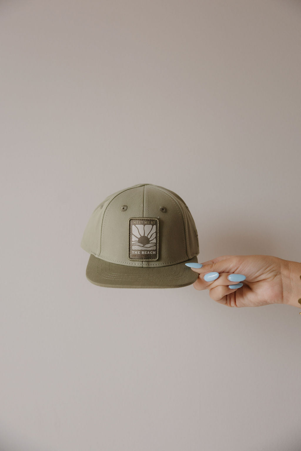 A hand with light blue nail polish holds the forever french baby Snapback | Meet Me at the Beach, a khaki green baby hat featuring a sun and wave patch, set against a plain, light-colored wall.