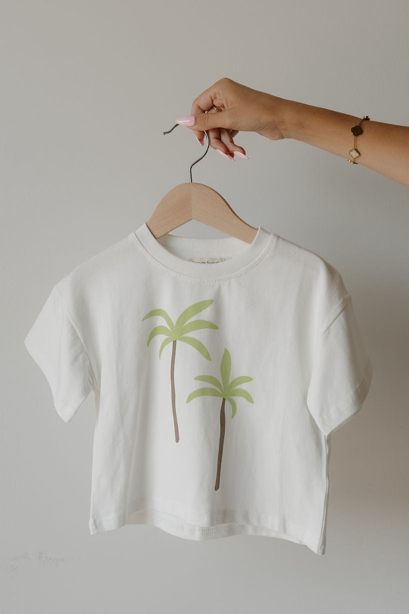 A hand holds a hanger with the forever french baby Child Cotton Tee | Palm, made from 100% cotton and featuring two green palm trees printed on the front, against a plain light background.