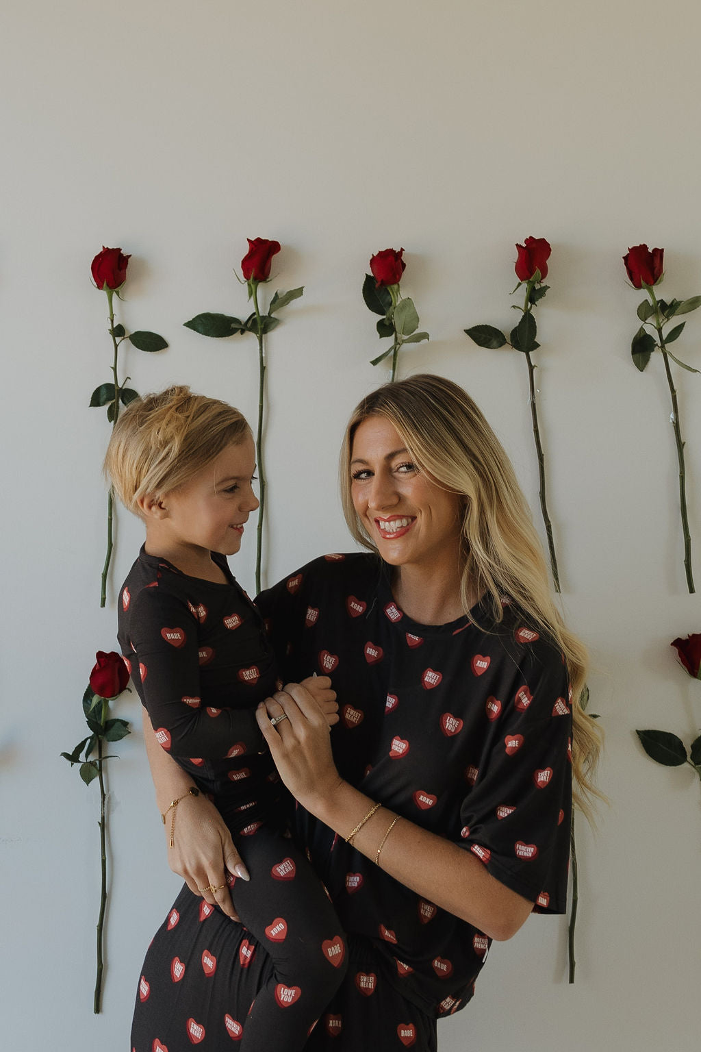 A smiling woman and child wear matching forever french baby Bamboo Two Piece Pajamas | Love Notes – Charcoal, featuring red hearts. Six red roses on the wall enhance their hypoallergenic kids sleepwear style.