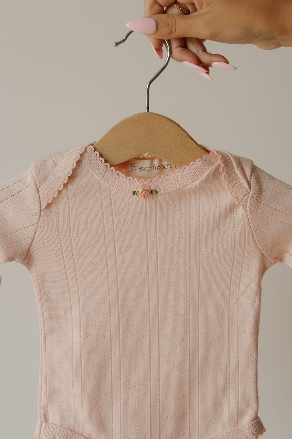 A hand with pink nails holds a hanger displaying the forever french baby Long Sleeve Snapsuit | Ballerina, a pale pink, long-sleeve pointelle cotton snapsuit featuring delicate knit details and a small floral accent at the neckline.