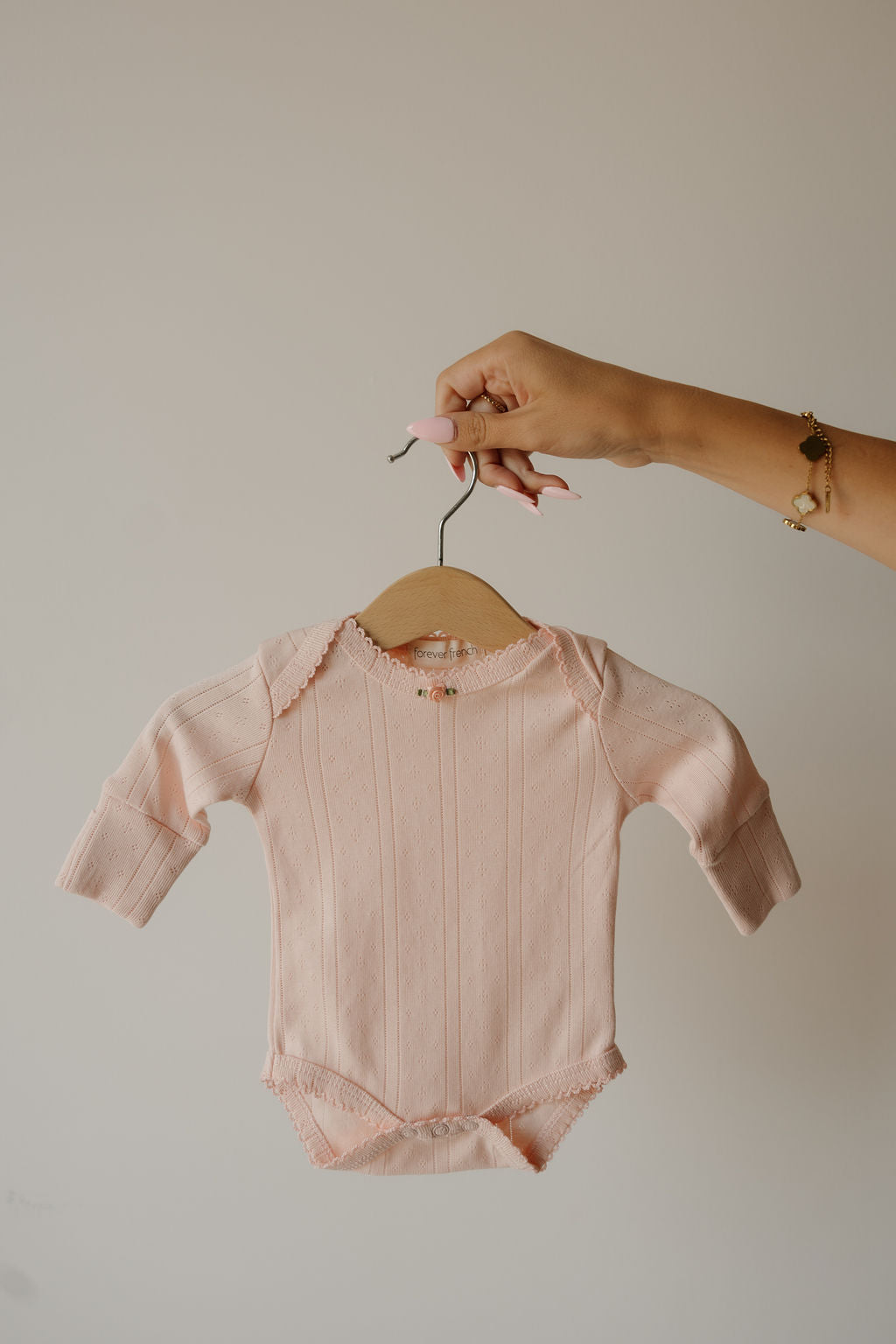 A hand holds the forever french baby Long Sleeve Snapsuit | Ballerina, a pale pink, long-sleeved cotton snapsuit, displayed against a plain, light-colored background.
