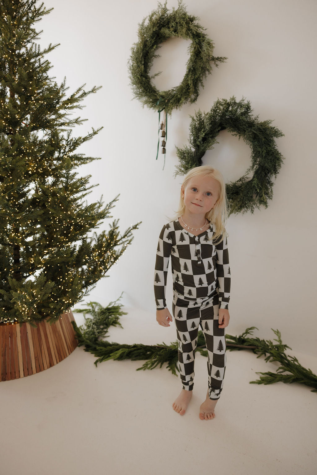 A young child in forever french baby’s Bamboo Two Piece Pajamas | Christmas Tree Checkerboard stands by a lit tree. Green wreaths hang on the white wall and garland decorates the floor, while their hypoallergenic bamboo pajamas ensure seasonal comfort.
