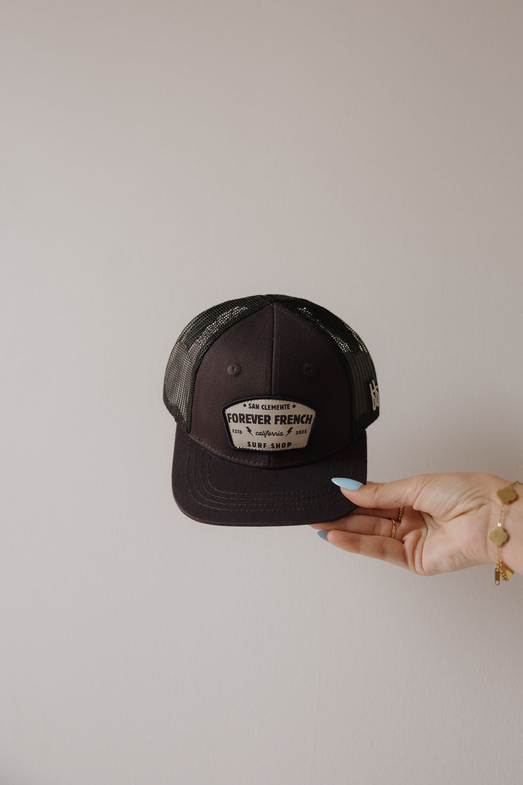 A hand with blue nail polish and a bracelet holds the "Snapback | ff Surf Shop" hat by forever french baby, featuring a patch, against a plain beige background.