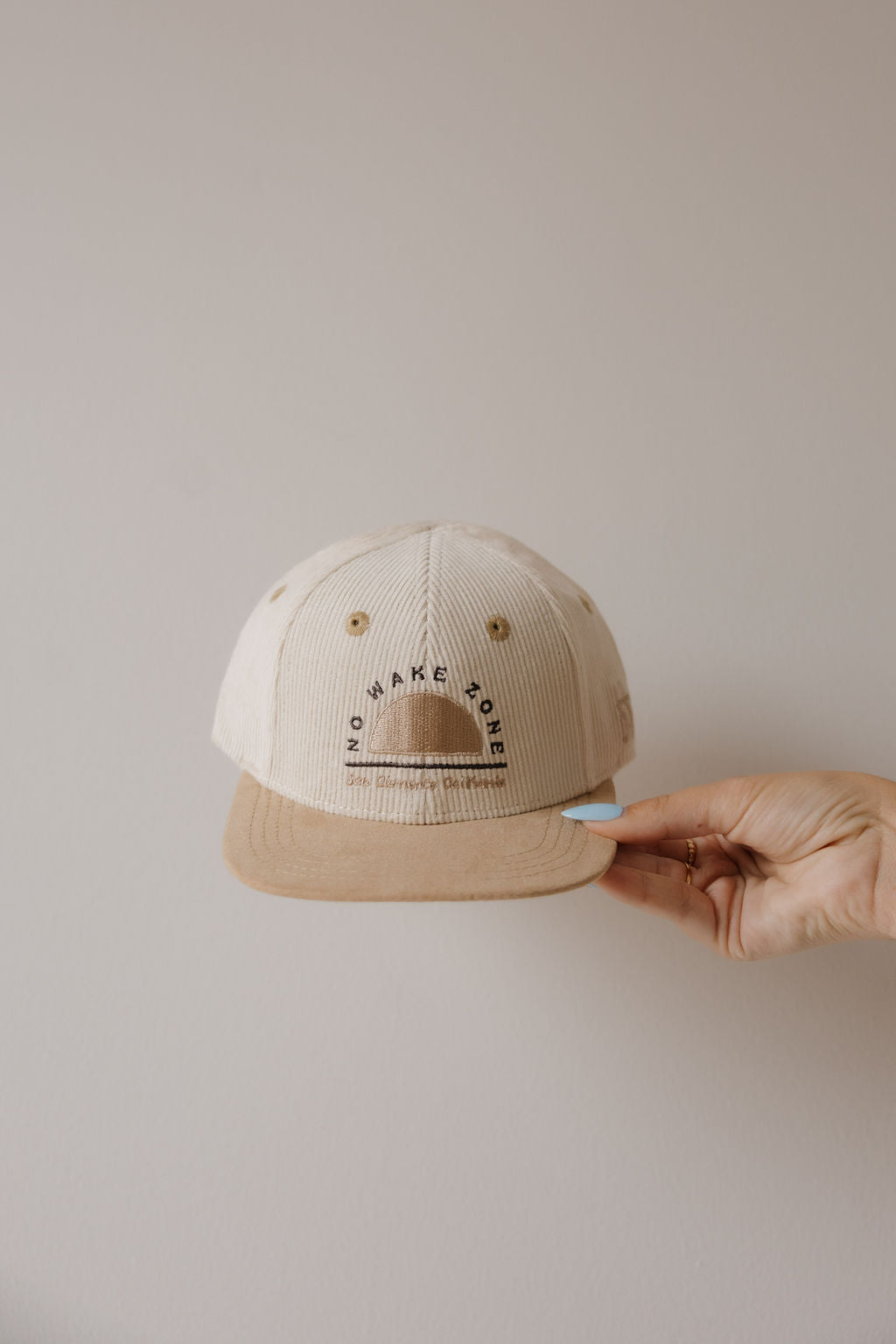 A hand with light blue nail polish holds the forever french baby Snapback | No Wake Zone cap, which features a beige corduroy crown, brown brim, simple sun graphic, and "ENJOY LAKE POWER" printed above in black letters.