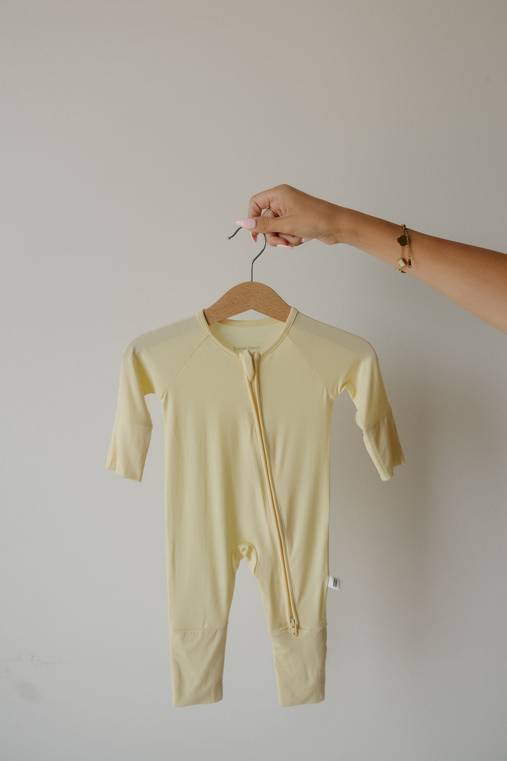 A hand holds a wooden hanger displaying the “Bamboo Zip Pajamas | Sunny” by forever french baby, a pale yellow long-sleeve baby onesie with a front zipper, set against a plain light background and highlighting its soft, hypoallergenic fabric that’s ideal for sensitive skin.