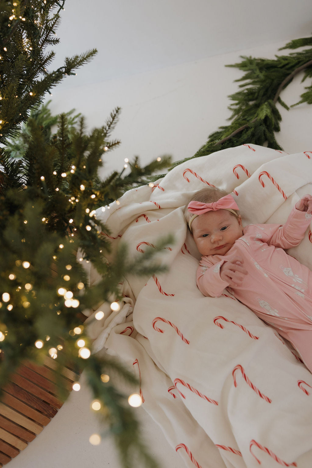 A baby in a pink onesie and headband lies on the forever french baby Muslin Quilt | Candy Cane, next to a decorated Christmas tree with lights and green garlands.