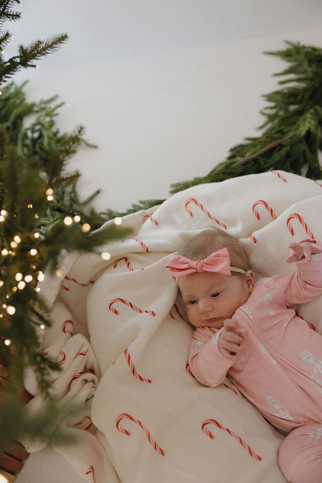 A baby in forever french baby Bamboo Zip Pajamas | Nutcracker and a pink headband lies on a white candy cane blanket beside a Christmas tree with twinkling lights and green branches.