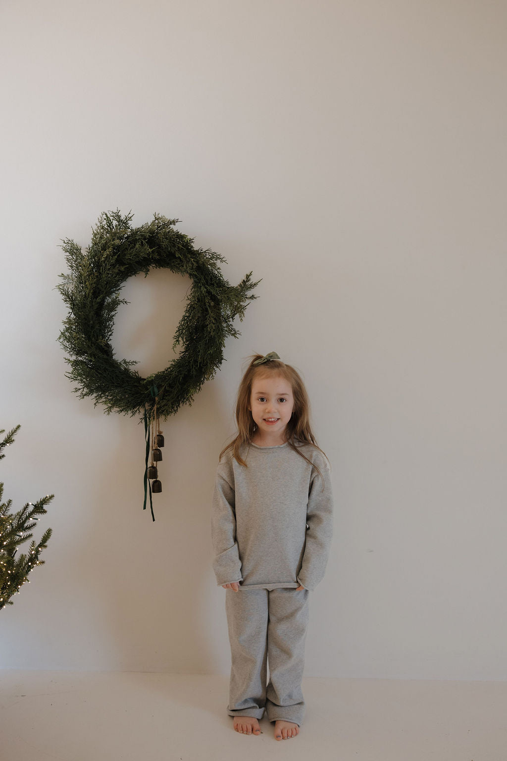 A young child wearing the forever french baby Knit Pant Set | Grey stands barefoot against a plain white wall, with a green holiday wreath and some greenery visible nearby.