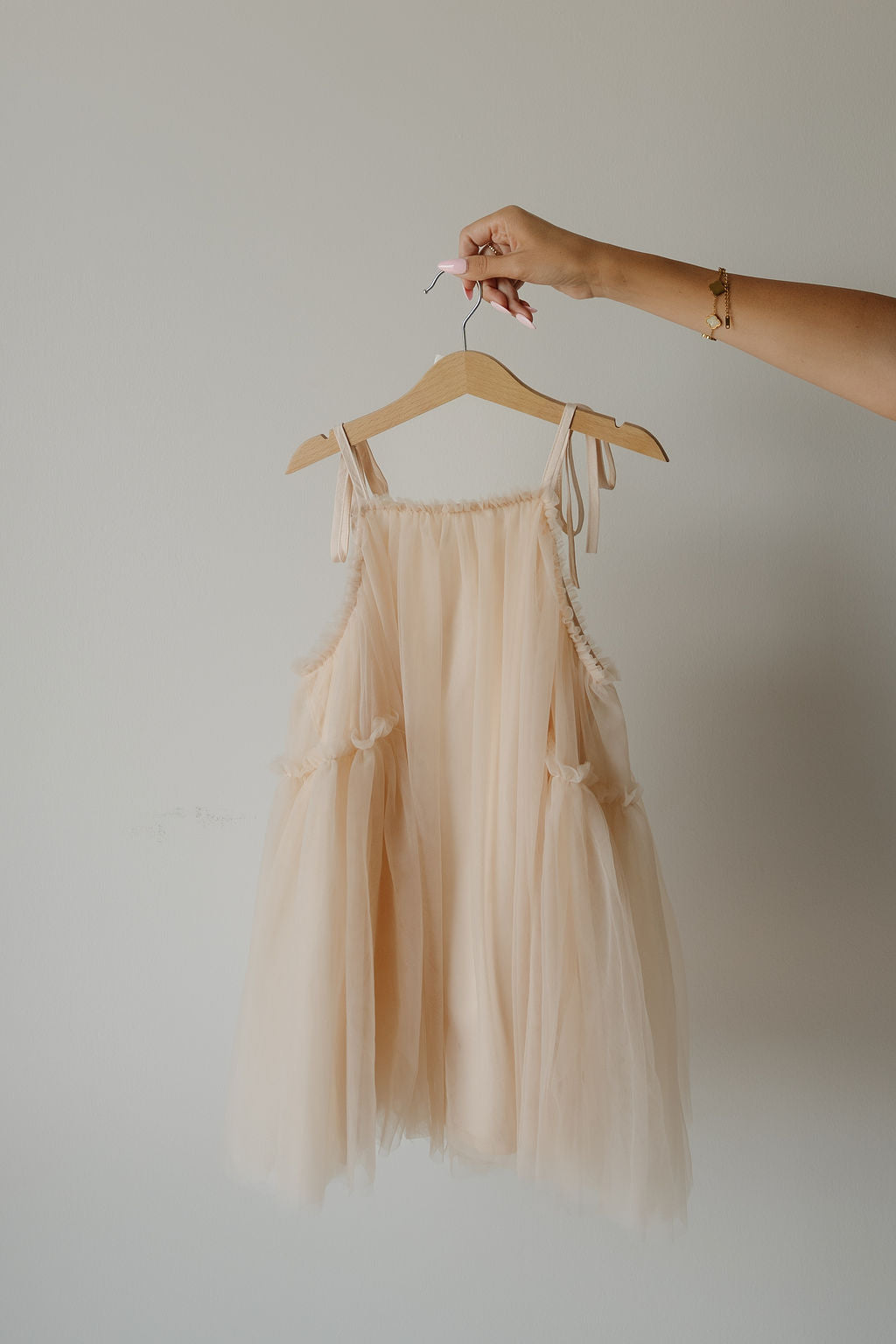 A hand holds a hanger with the forever french baby Tutu Dress in the Slipper shade, a light peach, sleeveless tulle tutu dress featuring delicate bow straps, set against a plain white background.