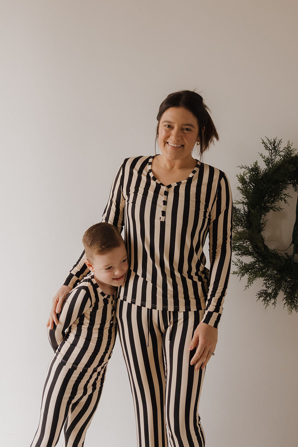 A woman and a young boy, both wearing matching Forever French Baby Women's Long Sleeve Bamboo Pajamas in Midnight Stripe, smile together in front of a white wall with a green wreath partially visible on the right.