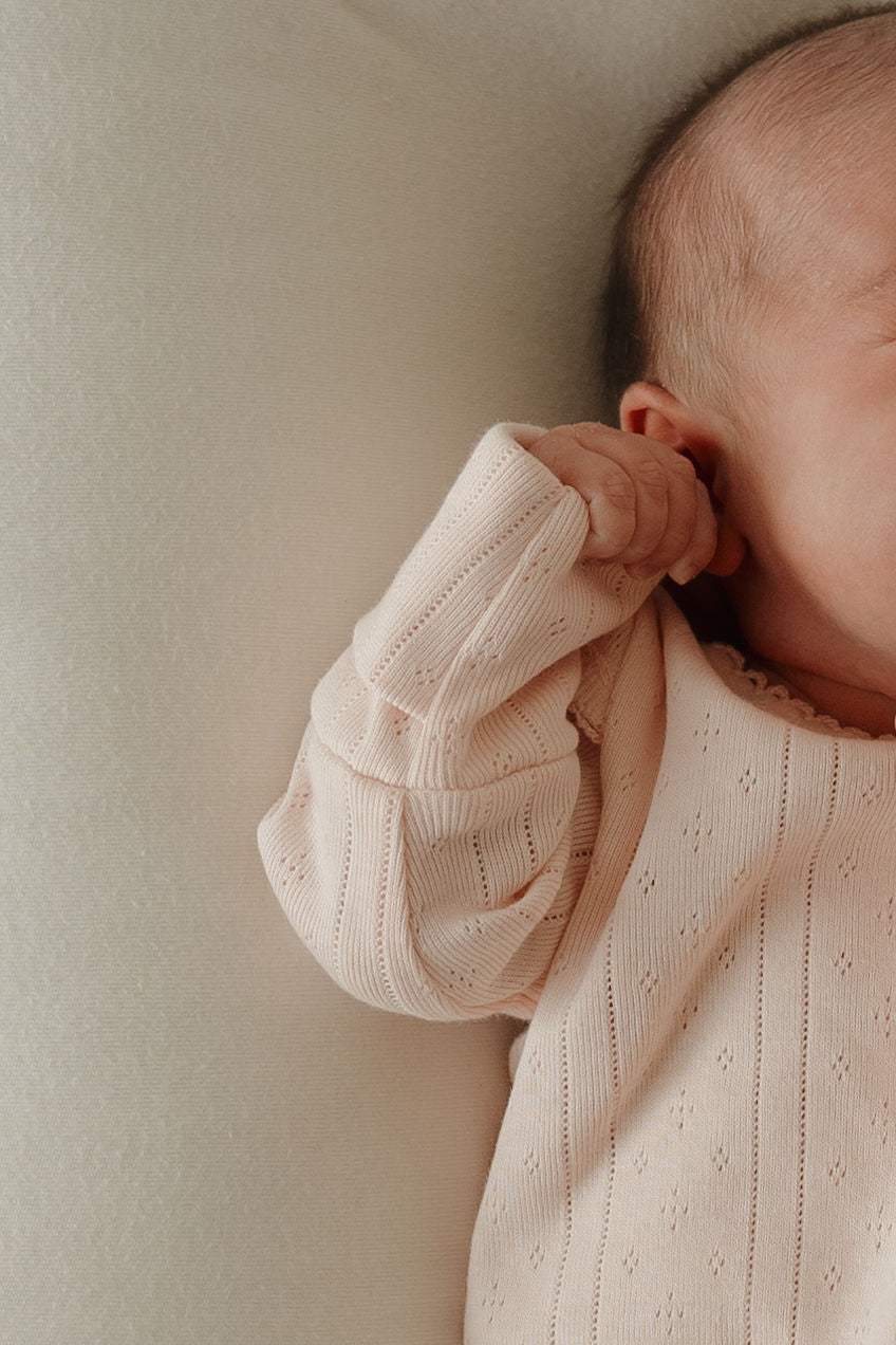 Close-up of a baby lying on a soft surface, wearing the forever french baby Long Sleeve Snapsuit | Ballerina in light pink pointelle cotton. The baby’s face is partially visible, with one hand near their ear.
